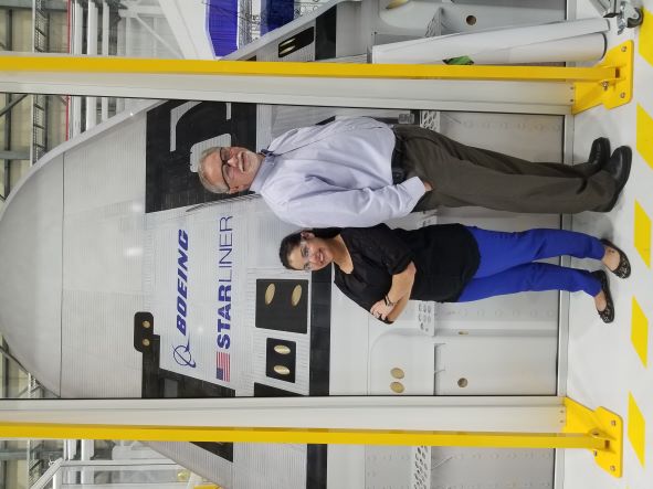 Melanie and Mike standing back-to-back in front of the Starliner. 