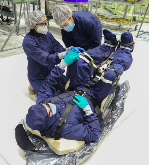 Two engineers are gathered around an employee sitting in the Starliner crew seat, making adjustments They are wearing blue jumpsuits, hair nets and face masks.