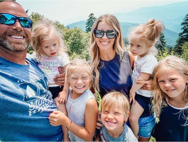 Rayni posing with her husband and their 5 children. They are standing in nature, smiling. 