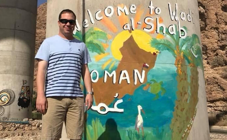 Elliot Riley standing in front of a bridge column with graffiti text reading Welcome to Wadi al-Shab, Oman
