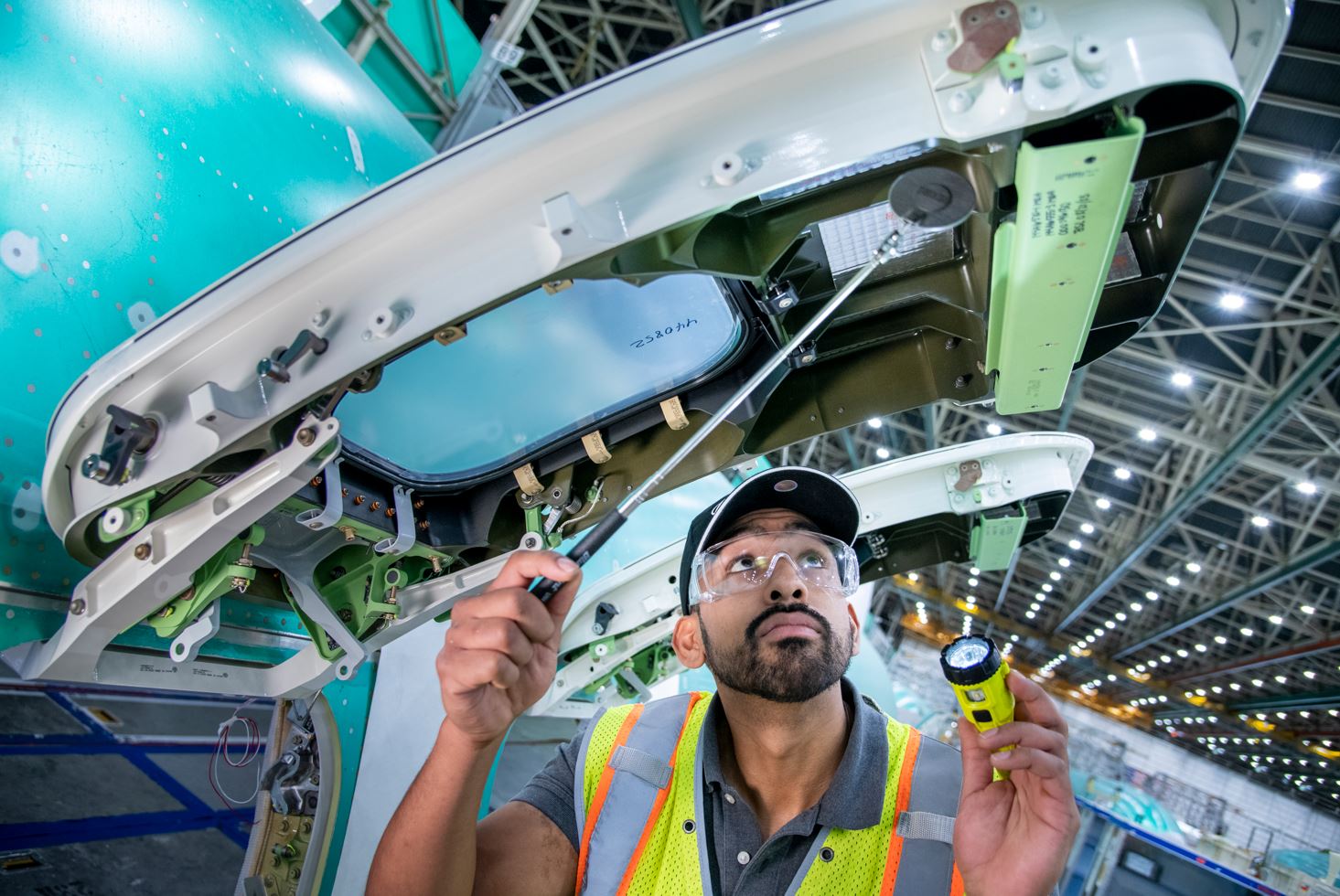 Joseph in a large factory setting, holding flashlight, looking upward into the overwing 