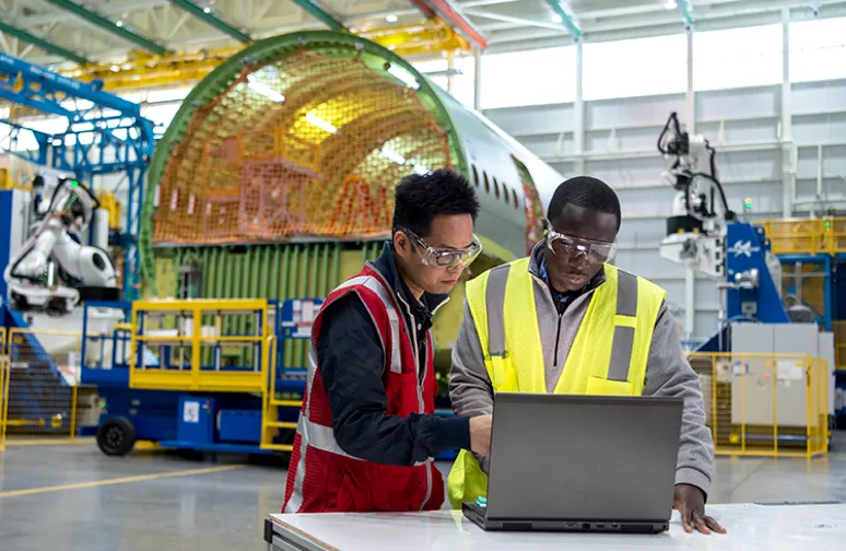two male engineers looking at a laptop together inside a large manufacturing facility