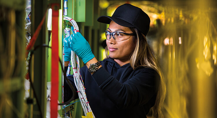Female employee wearing gloves and safety goggles, inspecting an electronics panel