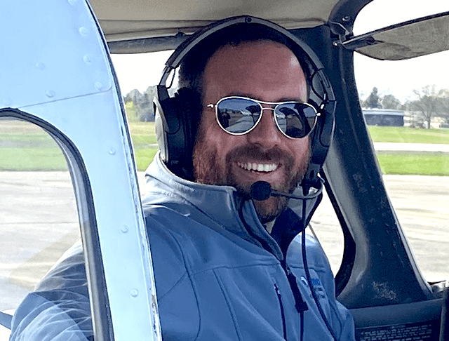 Dominic seated in a small aircraft with a headset on and aviator sunglasses, faces the camera smiling