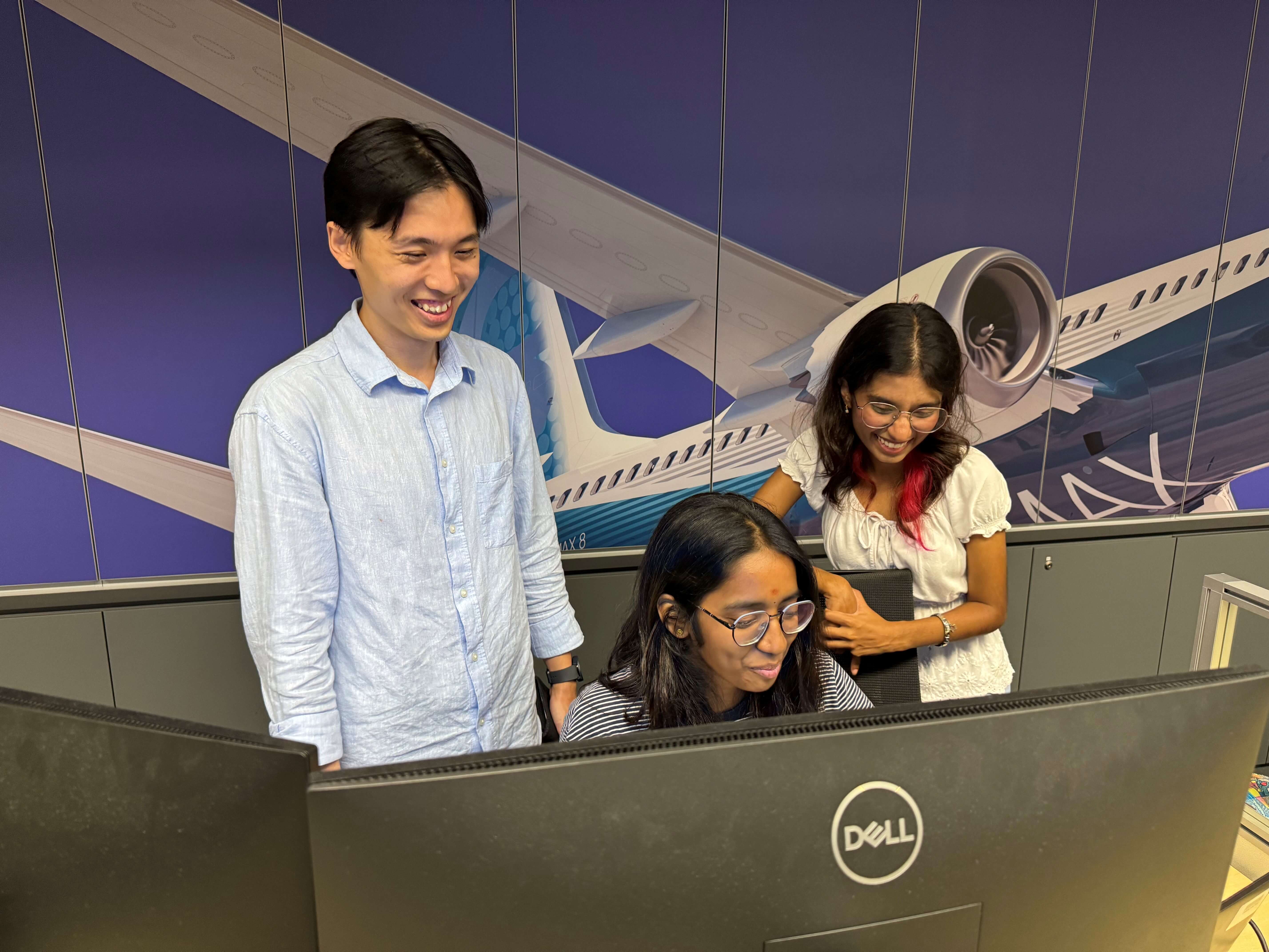 Three interns, one seated, two standing, huddle in front of dual monitors reviewing what’s on the screen 