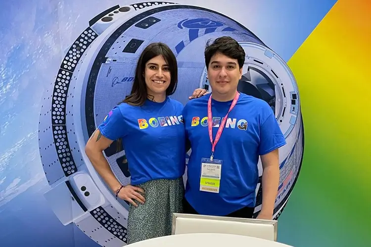2 young Boeing employees wearing Boeing t-shirts with rainbow colored text, standing in front of a table at an event