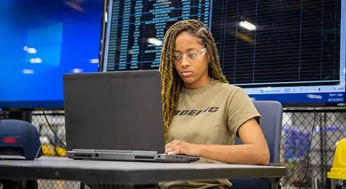 young black female programmer working on her laptop in front of a large panel of screens 