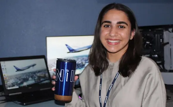 female youth sitting at her computer desk, holding up a travel mug and smiling