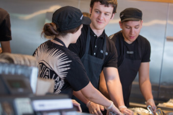 
Chipotle Restaurant Manager está capacitando a un nuevo miembro de la tripulación para construir cuencos y burritos para los huéspedes del restaurante.