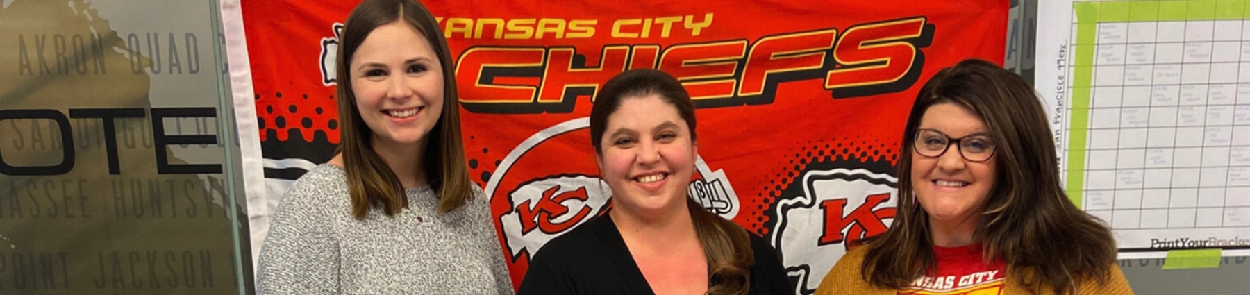 Three women smiling in front of Kansas City Chiefs poster