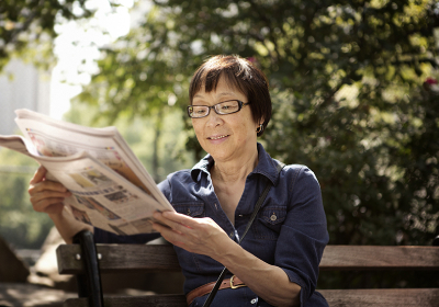 Woman with glasses reading newspaper on bench in park