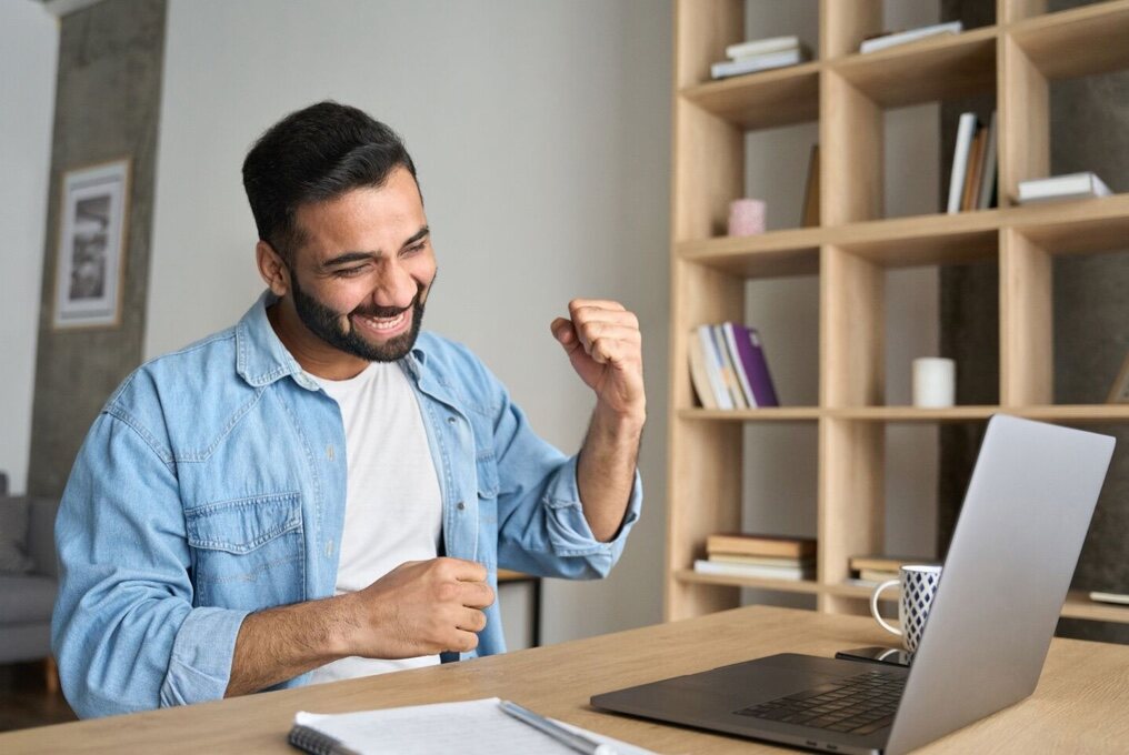 Um homem está sentado à sua mesa com um laptop, erguendo o punho com determinação.