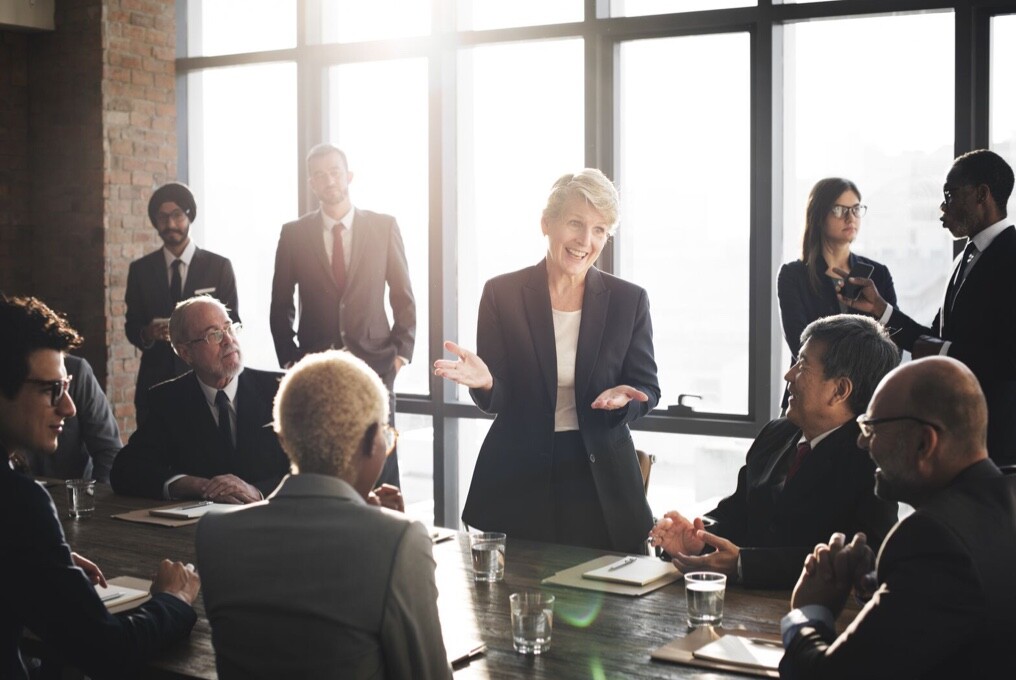 Eine Frau im Business-Anzug hält eine Präsentation vor einer Gruppe von Menschen.