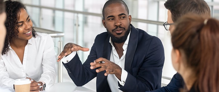 Male staff member in a blue suit and shirt speaking to his colleagues around a white table