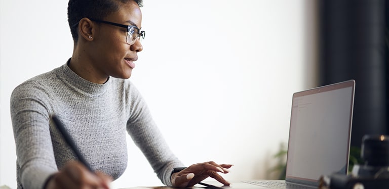 Woman with grey jumper and glasses with one hand on her laptop keyboard and the other hand making notes with a pen