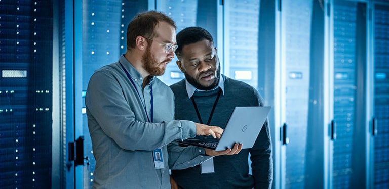 Two men in shirts and jumpers standing in a room full of server racks looking at a laptop screen