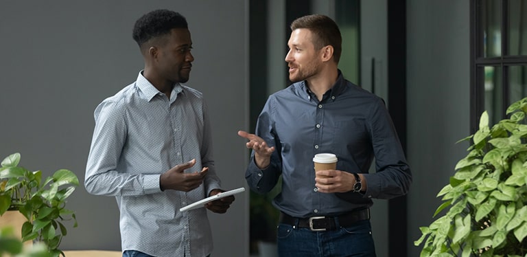 Two men in shirts in a discussion. One is holding a notebook, the other is holding a coffee cup