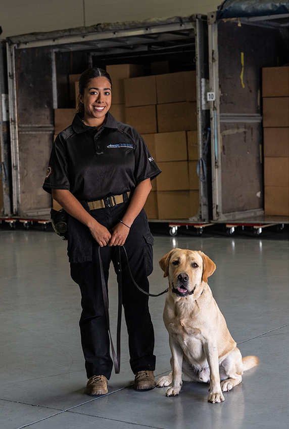 Uniformed K‑9 handler standing with a leashed dog inside a warehouse or loading facility, with large carts of stacked boxes visible in the background.