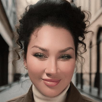 Person with curly hair standing in an outdoor urban walkway, wearing a light turtleneck and jacket.