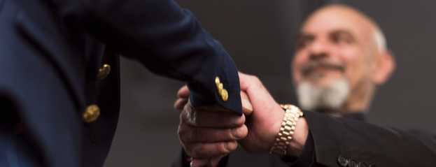 Two professionals shake hands in a close-up view, with one wearing a dark suit jacket with gold buttons and the other wearing a watch, conveying a formal agreement or professional partnership.