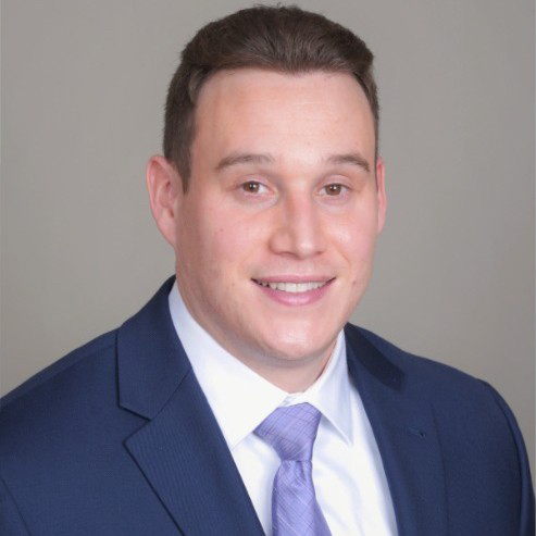 A person wearing a navy suit jacket, white dress shirt, and light purple tie poses for a professional headshot against a plain background.