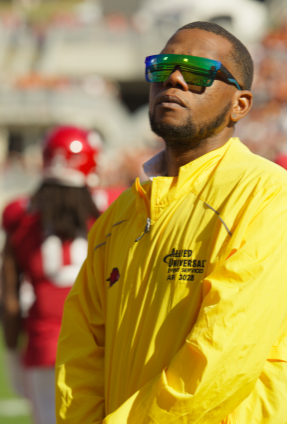 An event staff member wearing a bright yellow jacket stands on a sports field during a live game, with players and a crowded stadium visible in the background.