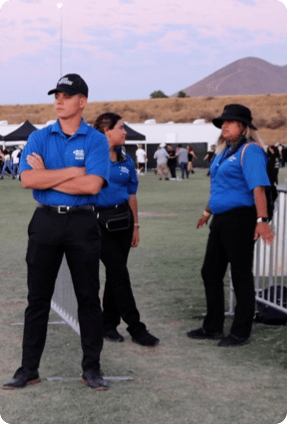 Event staff in matching blue uniforms stand on a grassy field near temporary barriers, monitoring an outdoor gathering with tents, attendees, and a mountain landscape in the background.