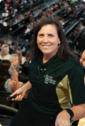 An event staff member wearing an Allied Universal polo shirt sits in an arena with a crowd in the background.
