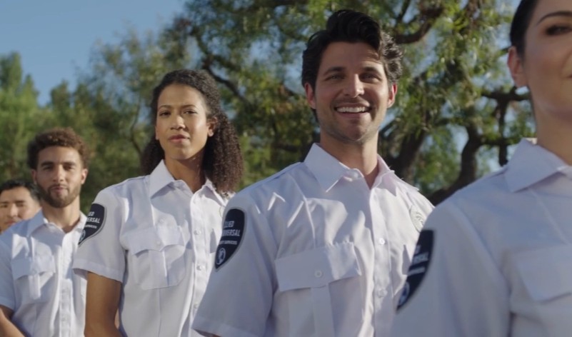 A diverse group of uniformed security professionals standing in formation outdoors, wearing white short-sleeve shirts with shoulder patches, with trees and daylight in the background.
