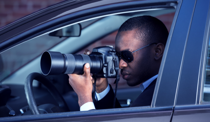 A person sitting inside a car aims a large telephoto camera lens out the open window, appearing to take photographs or observe a subject from the vehicle.