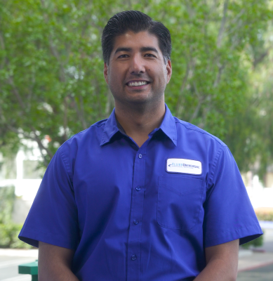 A person wearing a blue short-sleeve work shirt with a name badge stands outdoors in front of green trees, with hands relaxed at their sides.