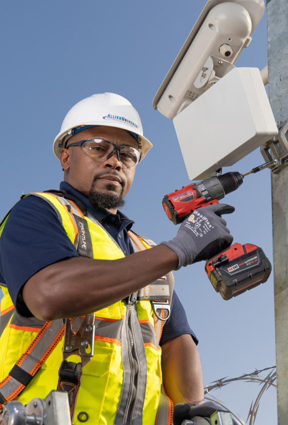 Utility worker wearing a hard hat and high‑visibility safety vest uses a power drill to install or service equipment mounted on a utility pole against a clear blue sky.