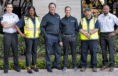 A group of security and safety professionals stand outdoors in a landscaped area, wearing a mix of uniforms, safety vests, and work attire, posed together as a team in front of a building entrance.