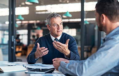 Two professionals sit across from each other at a table in a modern workspace, engaged in discussion, with one gesturing while speaking and documents spread out between them.