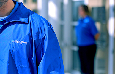 A staff member wearing a blue branded jacket stands inside a secure facility or workplace corridor, with another uniformed employee visible in the background near glass doors and interior lighting.