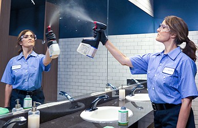 A custodial worker in a blue uniform sprays disinfectant onto a restroom mirror and sink, with cleaning supplies arranged on the counter in a clean, well‑lit facility.