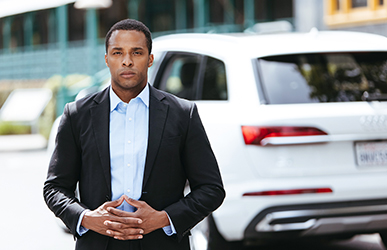 A professional wearing a dark blazer over a light blue shirt stands outdoors with hands clasped, positioned in front of a white SUV in an urban setting with modern buildings in the background.