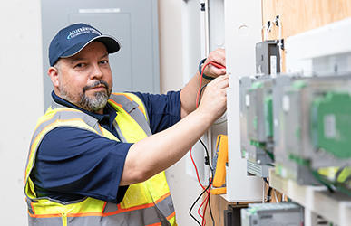 A utility worker wearing a high‑visibility safety vest and hard hat adjusts wiring inside an electrical control panel, using tools in an indoor industrial setting with equipment mounted along the wall.