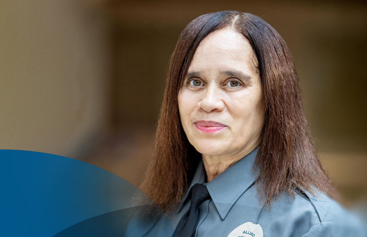 A person wearing a blue security uniform shirt and dark tie, standing indoors against a softly blurred background, with long straight hair and a visible name badge on the chest.