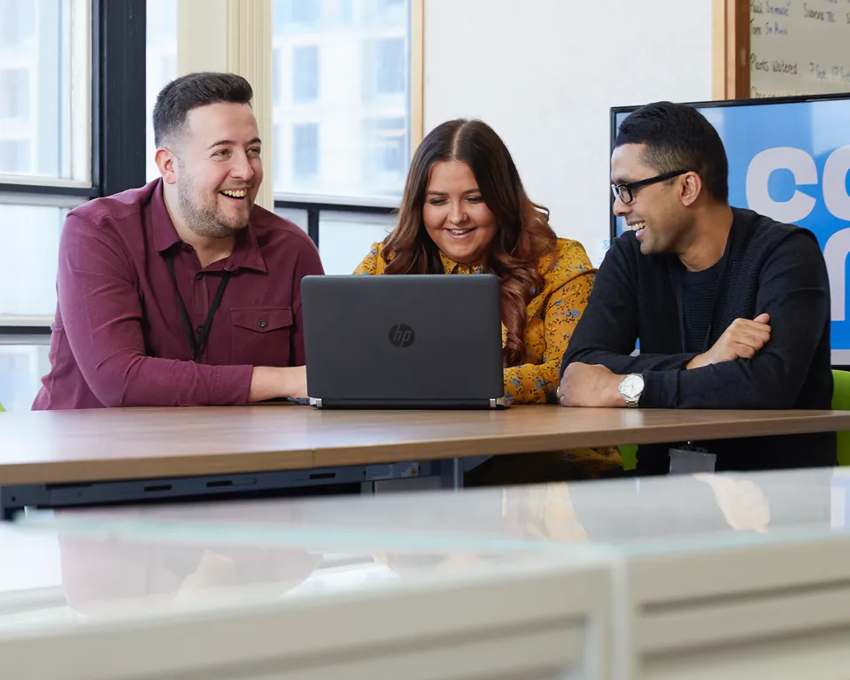 Three colleagues collaborate around a table with a laptop in a modern office setting