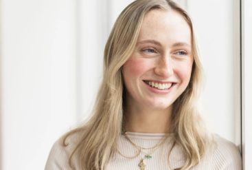Smiling person standing indoors near a white wall and a plant