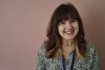 Smiling woman with bangs standing against pink wall.