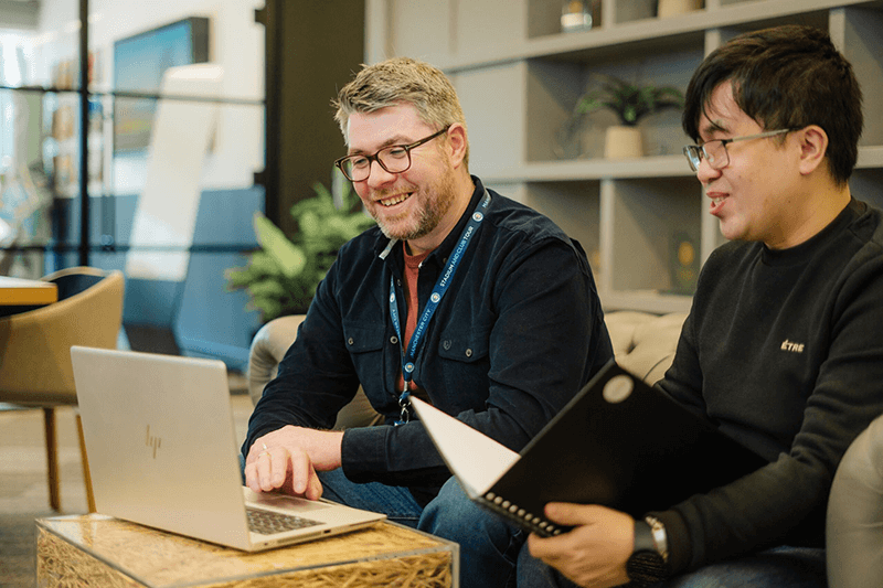 Two people collaborate in a modern office, working with a laptop and notebook at a shared desk.