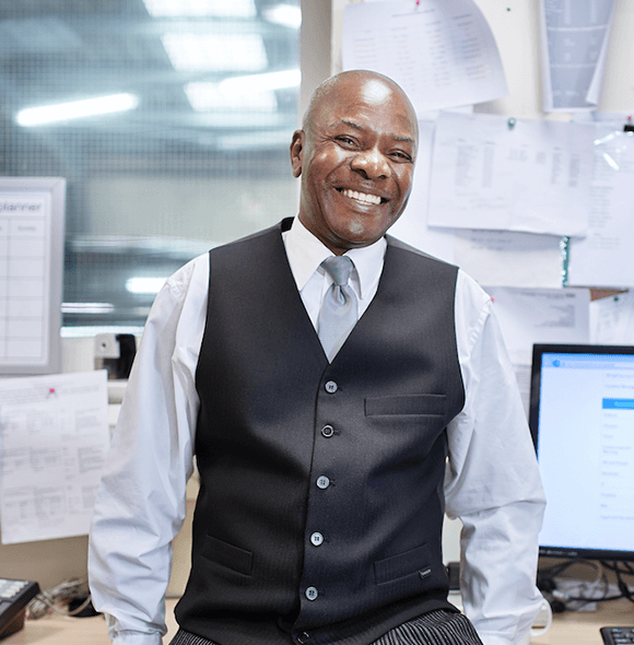 Individual in a white shirt, tie, and dark vest sits in an office with papers pinned to the wall and a computer screen visible to the right.