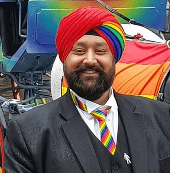 Individual in a black suit, white shirt, and rainbow turbant and tie stands near a colorful structure