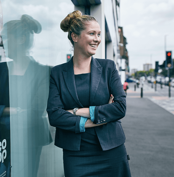 Professional individual in a business suit stands with arms crossed, leaning against a glass window on a city street