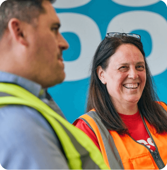 Two professionals in safety vests converse in front of a blue wall with the Co-op logo