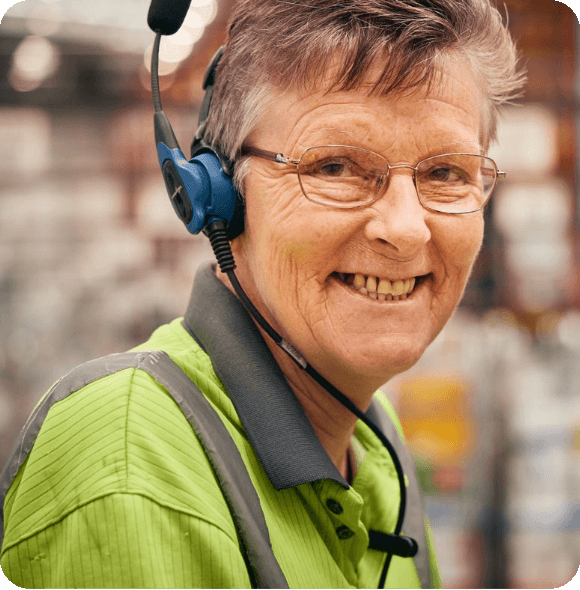 Warehouse worker in a safety vest and headset stands in an industrial environment, surrounded by shelves and equipment