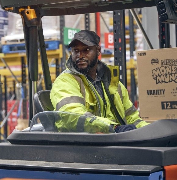Person in a high-visibility jacket and cap operates a forklift in a warehouse