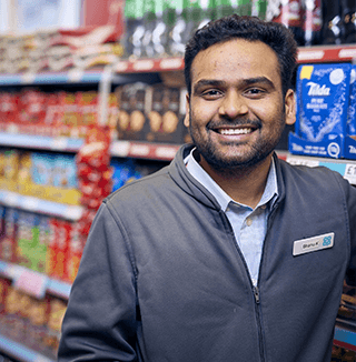 A smiling employee stands in a retail setting, surrounded by shelves stocked with various products.